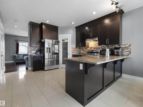 Modern kitchen featuring dark wood cabinetry, stainless steel appliances, a multi-tone tile backsplash, and a large central island with an integrated sink and light granite-style countertop - 5192 1B Ave Sw, Edmonton, AB - Indoor Photo Showing Kitchen With Upgraded Kitchen