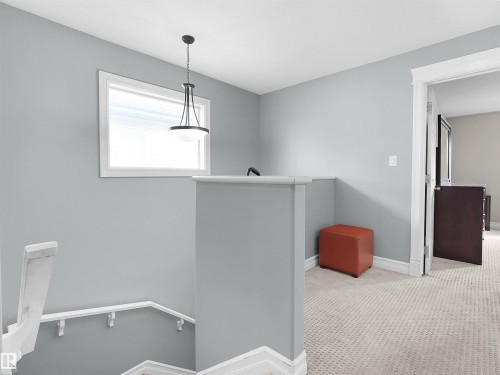 Carpeted upper landing featuring a modern pendant light fixture, a window with white trim, and a white handrail leading downstairs - 5192 1B Ave Sw, Edmonton, AB - Indoor Photo Showing Other Room