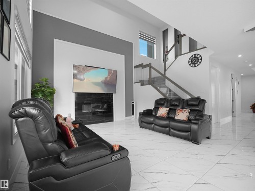 Open-concept living space with large format white tile flooring, a contemporary fireplace feature wall with a mounted television, a modern staircase with glass railing, and recessed lighting throughout - 1208 25 St Nw, Edmonton, AB - Indoor