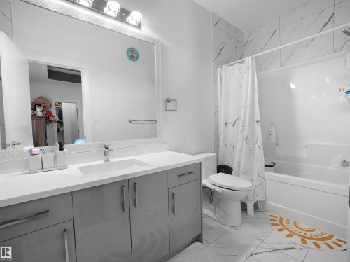 Modern bathroom featuring white tile flooring with veining, a gray vanity with an integrated sink and chrome fixtures, a large frameless mirror with overhead lighting, and a combination tub and shower with tiled surround - 1208 25 St Nw, Edmonton, AB - Indoor Photo Showing Bathroom