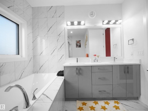 Contemporary bathroom featuring an integrated bathtub with chrome fixtures, a double vanity with a light gray finish, a large mirror, and white marble-look wall and floor tiling - 1208 25 St Nw, Edmonton, AB - Indoor Photo Showing Bathroom