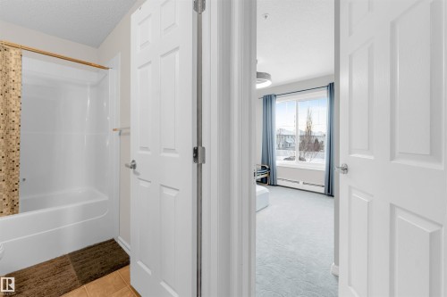 Bright interior featuring a white bathtub with shower, ceramic tile flooring, and a window with blue drapes - 201 4403 23 Street, Edmonton, AB - Indoor Photo Showing Other Room
