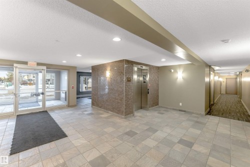 Spacious entrance lobby featuring large format tile flooring, a textured accent wall, stainless steel elevator, recessed lighting, and glass entry doors - 201 4403 23 Street, Edmonton, AB - Indoor Photo Showing Other Room