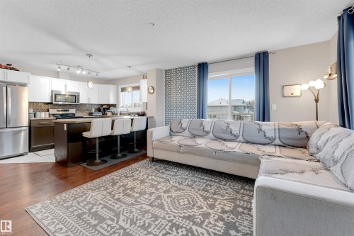 Open-concept living space featuring wood-finish flooring, a kitchen island with seating, stainless steel appliances, white upper cabinetry, and a patterned accent wall - 201 4403 23 Street, Edmonton, AB - Indoor Photo Showing Living Room