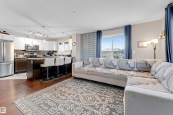 Open-concept living space featuring wood-finish flooring, a kitchen island with seating, stainless steel appliances, white upper cabinetry, and a patterned accent wall - 