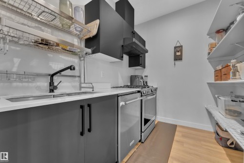 Contemporary kitchen featuring dark gray lower cabinetry with matte black hardware, a stainless steel gas range with coordinating range hood, a stainless steel dishwasher, and light wood-finish flooring - 5722 Kootook Way, Edmonton, AB - Indoor Photo Showing Kitchen With Double Sink