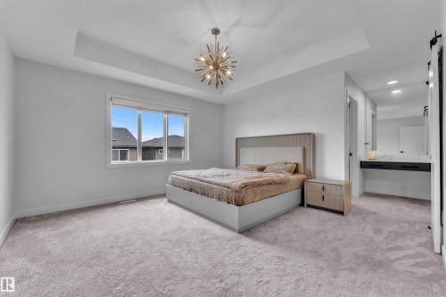 Carpeted bedroom featuring a tray ceiling with a contemporary light fixture, large window, and a built-in vanity with a countertop and undermount sink - 5722 Kootook Way, Edmonton, AB - Indoor Photo Showing Bedroom