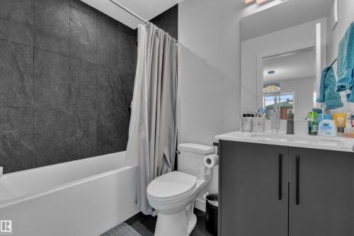 Contemporary bathroom featuring a white bathtub, dark gray textured wall tile, a sleek vanity with a light-toned countertop, a rectangular mirror, and dark-toned cabinetry with vertical hardware - 5722 Kootook Way, Edmonton, AB - Indoor Photo Showing Bathroom