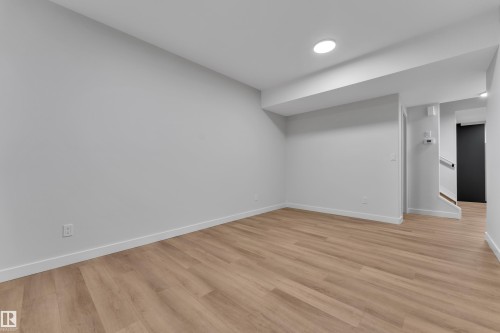 Spacious room featuring wood-finish flooring, recessed ceiling lighting, light gray walls, white baseboards, and a contemporary staircase with a dark door - 5722 Kootook Way, Edmonton, AB - Indoor Photo Showing Other Room