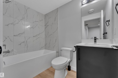 Contemporary bathroom featuring a white bathtub with large-format marble-look wall tiles, a dark gray vanity with a white countertop and undermount sink, a wall-mounted mirror, and wood-finish flooring - 5722 Kootook Way, Edmonton, AB - Indoor Photo Showing Bathroom