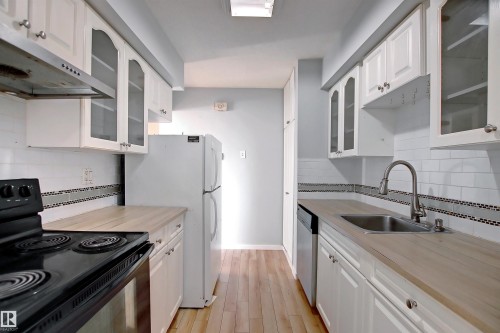Galley kitchen featuring light wood-finish countertops, white cabinetry with some glass-front doors, white subway tile backsplash with a mosaic accent, stainless steel sink, and wood-finish flooring - 11 14120 80 Street, Edmonton, AB - Indoor Photo Showing Kitchen