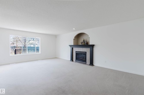Spacious room featuring a fireplace with a dark wood mantel and tile surround, an arched niche above the fireplace, a large window with white framing, and light-colored carpeting - 1951 120 Street, Edmonton, AB - Indoor Photo Showing Living Room With Fireplace