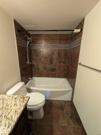 Bathroom featuring a tub/shower combination with brown tile surround and decorative accent band, a white toilet, and a vanity with a granite-style countertop - 