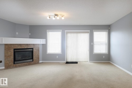 Carpeted living space featuring a tile-surround fireplace with a white mantel, a sliding glass door, and two windows with horizontal blinds - 9071 Scott Crescent, Edmonton, AB - Indoor Photo Showing Living Room With Fireplace