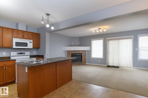 Kitchen and living area featuring a central island with dual sinks, wood-finish cabinetry, a white appliance suite, a fireplace with a mantel, and a sliding glass door - 9071 Scott Crescent, Edmonton, AB - Indoor Photo Showing Kitchen With Fireplace