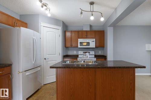 Kitchen featuring a central island with a double basin sink, rich wood-finish cabinetry, white appliances, and track lighting - 9071 Scott Crescent, Edmonton, AB - Indoor Photo Showing Kitchen With Double Sink