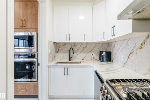 Modern kitchen featuring white cabinetry, a built-in stainless steel microwave and oven, a black sink faucet, a cooktop, and light-toned countertops with a complementary backsplash - 219 28 Street, Edmonton, AB - Indoor Photo Showing Kitchen With Double Sink