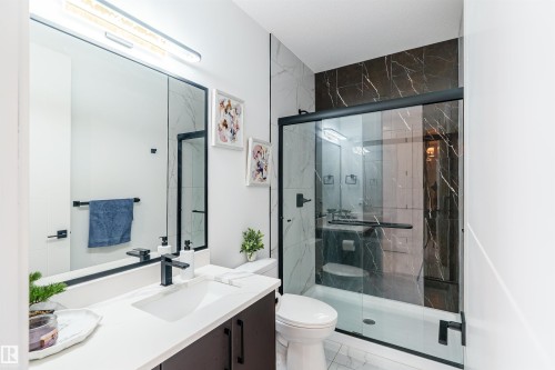 Modern bathroom featuring a single vanity with a white countertop and dark cabinetry, a frameless mirror with integrated lighting, a toilet, and a glass-enclosed shower with contrasting dark and light marble-look tile - 219 28 Street, Edmonton, AB - Indoor Photo Showing Bathroom