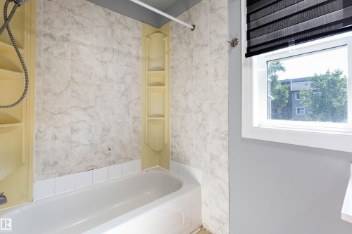 White porcelain bathtub with integrated shelving, surrounded by beige patterned wall tiles and a white window frame with dark roller blinds - 547 Dunluce Road, Edmonton, AB - Indoor Photo Showing Bathroom