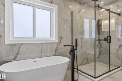 Modern bathroom featuring a freestanding soaking tub with a matte black floor-mounted faucet, a glass-enclosed shower with matte black fixtures, full-height veined tile walls, and a large window with white trim - 