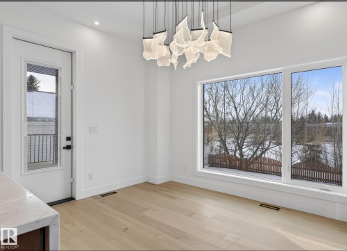 Expansive windows with a winter tree line backdrop, featuring a contemporary chandelier, light wood-finish flooring, and a white paneled door with matte black hardware - 5820 110 Street, Edmonton, AB - Indoor Photo Showing Other Room