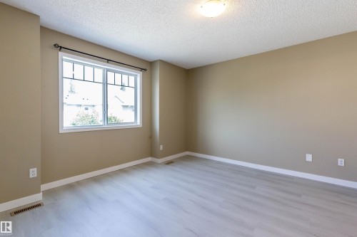Spacious room featuring light wood-finish flooring, neutral wall tones, white baseboards, and a large window with divided lites and a black decorative curtain rod - #116 5001 62 St, Beaumont, AB - Indoor Photo Showing Other Room