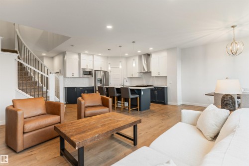 Open-concept living space featuring wood-finish flooring, a contemporary kitchen with a large island and stainless steel appliances, and a curved staircase with metal spindles - 6003 Edmonds Place Nw, Edmonton, AB - Indoor Photo Showing Living Room