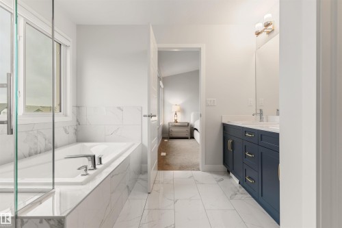 Contemporary bathroom featuring a built-in tub with tile surround, a spacious double vanity with a dark blue cabinet, chrome fixtures, and large format tile flooring - 6003 Edmonds Place Nw, Edmonton, AB - Indoor Photo Showing Bathroom