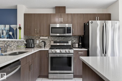 Kitchen featuring wood-finish cabinetry, stainless steel appliances, a mosaic tile backsplash, and light-toned countertops - 16 7503 Getty Gate, Edmonton, AB - Indoor Photo Showing Kitchen With Stainless Steel Kitchen With Double Sink With Upgraded Kitchen