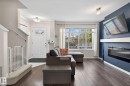 Open concept living area featuring dark wood-finish flooring, a wall-mounted electric fireplace with a white mantel, and a large front window - 16 7503 Getty Gate, Edmonton, AB  - Indoor Photo Showing Living Room With Fireplace 