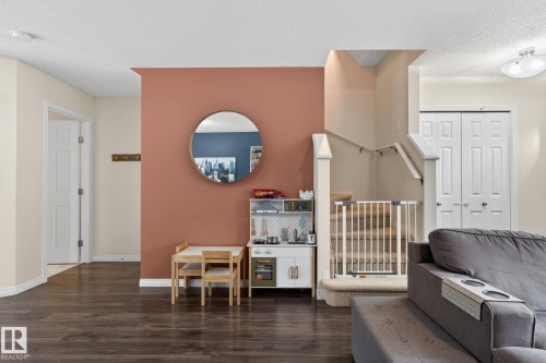 Open concept living space featuring wood-finish flooring, a contemporary accent wall, a circular wall mirror, a carpeted staircase with a railing, and white bi-fold closet doors - 16 7503 Getty Gate, Edmonton, AB - Indoor