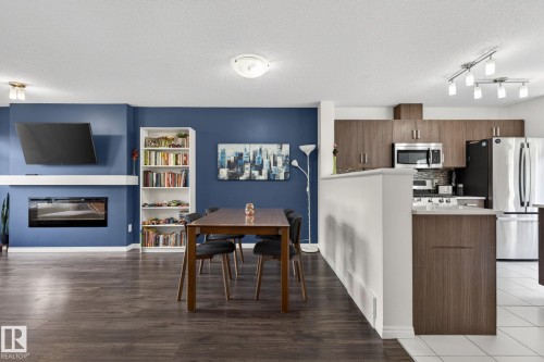 Spacious open-concept living area featuring wood-finish flooring, a built-in electric fireplace with white mantel, a white built-in bookshelf, and a kitchen with stainless steel appliances, dark wood-finish cabinetry, and track lighting - 16 7503 Getty Gate, Edmonton, AB - Indoor Photo Showing Kitchen