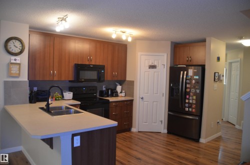Contemporary kitchen featuring wood-finish cabinetry, light-toned countertops, stainless steel double basin sink with matte black faucet, and coordinating black appliances - 1412 152, Edmonton, AB - Indoor Photo Showing Kitchen With Double Sink