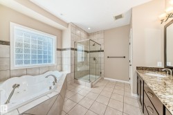 Bathroom featuring a spacious jetted tub with tiled surround, a glass-enclosed shower with tile walls, a vanity with a granite-style countertop and integrated sink, dark wood-finish cabinetry, and a large privacy glass block window - 