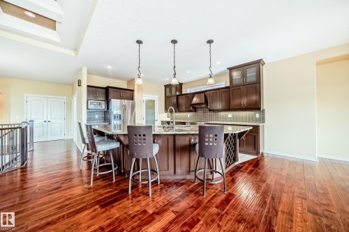 Open-concept kitchen featuring dark wood cabinetry, stainless steel appliances, a tile backsplash, a large island with a breakfast bar, and wood-finish flooring - 527 Callaghan Point(E), Edmonton, AB - Indoor