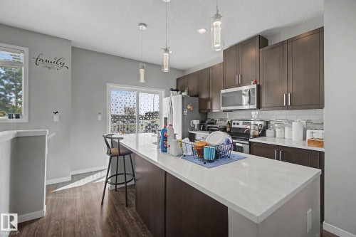 Modern kitchen featuring a large island with a light-toned countertop, dark wood-finish cabinetry, stainless steel appliances, and a tiled backsplash - 17 320 Secord Boulevard, Edmonton, AB - Indoor Photo Showing Kitchen