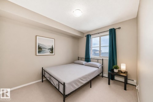 Bedroom featuring neutral tone walls, carpeted flooring, a ceiling-mounted light fixture, and a window with teal curtains - 404 12025 22 Avenue, Edmonton, AB - Indoor Photo Showing Bedroom
