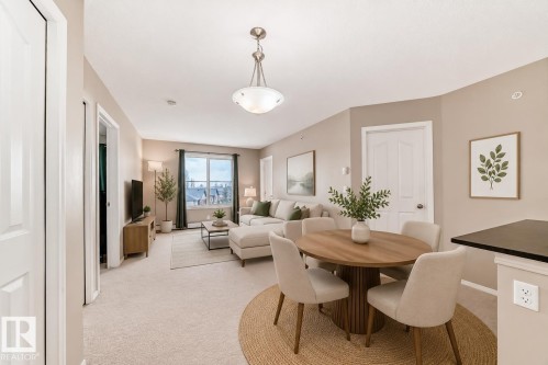Open-concept living area with neutral carpeting, a large window, a dining area featuring a pendant light fixture, and a built-in breakfast bar with a dark countertop - 404 12025 22 Avenue, Edmonton, AB - Indoor Photo Showing Dining Room