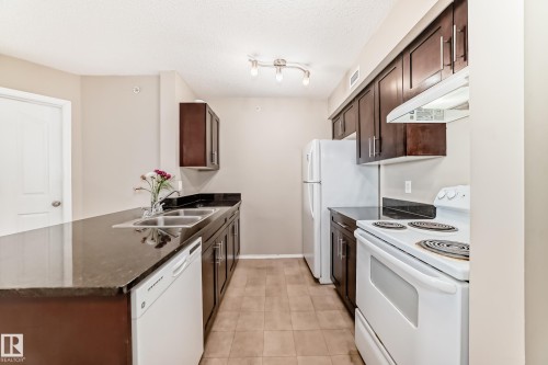 Kitchen featuring dark wood-finish cabinetry, dark countertops, and a full suite of white appliances including a refrigerator, range, and dishwasher - 404 12025 22 Avenue, Edmonton, AB - Indoor Photo Showing Kitchen With Double Sink