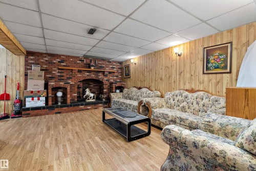 Spacious recreation room featuring brick fireplace with a raised hearth, wood-finish flooring, wood paneling, and a dropped ceiling with integrated lighting - 15907 101 Street, Edmonton, AB - Indoor With Fireplace