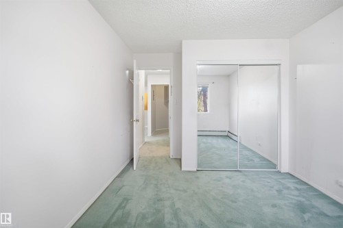 Light-colored carpeted room featuring a mirrored sliding closet door, white walls, and a window providing natural illumination - 124 10511 42 Avenue, Edmonton, AB 