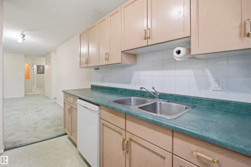 Kitchen area featuring wood-finish cabinetry with brass-tone hardware, a double-basin stainless steel sink, and a white tiled backsplash - 124 10511 42 Avenue, Edmonton, AB 