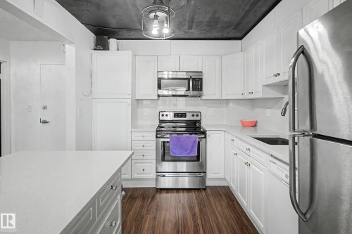 Modern kitchen featuring white shaker cabinetry, stainless steel appliances, wood-finish flooring, a kitchen island with a light-toned countertop, and an exposed ceiling - 1205 10045 118 Street, Edmonton, AB - Indoor Photo Showing Kitchen With Upgraded Kitchen