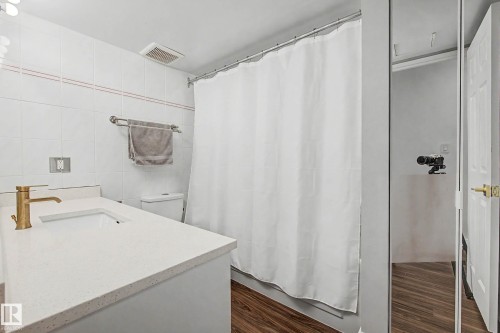 Bathroom featuring a white vanity with an integrated rectangular sink, a gold-finish faucet, white wall tiling with an accent stripe, and wood-finish flooring - 1205 10045 118 Street, Edmonton, AB - Indoor Photo Showing Bathroom