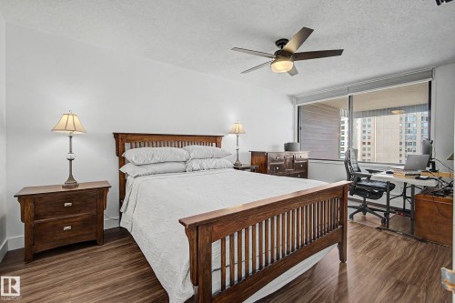 Spacious room featuring wood-finish flooring, a contemporary ceiling fan with integrated lighting, and a large window offering urban scenery - 1205 10045 118 Street, Edmonton, AB - Indoor Photo Showing Bedroom