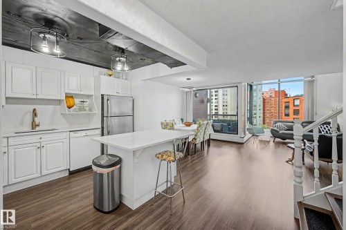 Open-concept living area featuring wood-finish flooring, a contemporary kitchen with white cabinetry and stainless steel appliances, a kitchen island with seating, a dedicated dining area, and large windows providing urban scenery - 1205 10045 118 Street, Edmonton, AB - Indoor Photo Showing Kitchen