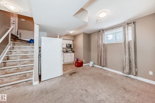 Lower level with carpeted flooring, featuring a window with privacy curtains, a compact kitchen area with white cabinetry, and a staircase with wood-finish treads and white risers - 1 12833 123 Street, Edmonton, AB - Indoor Photo Showing Other Room