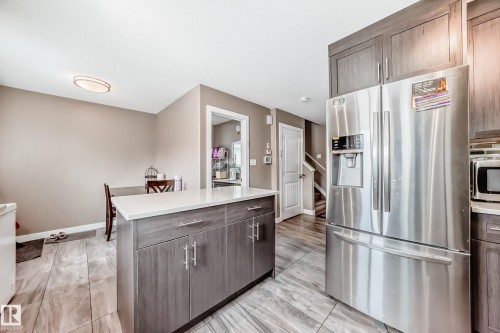 Kitchen featuring a central island with a light-toned countertop, wood-finish cabinetry, stainless steel French door refrigerator, and wood-look tile flooring - 1 12833 123 Street, Edmonton, AB - Indoor Photo Showing Kitchen