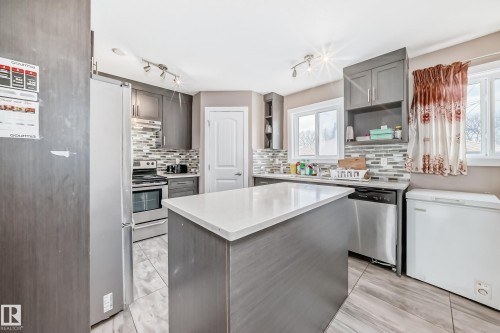 Kitchen featuring a central island with a solid surface countertop, wood-finish flooring, full-height cabinetry, stainless steel appliances, and a tiled backsplash - 1 12833 123 Street, Edmonton, AB - Indoor Photo Showing Kitchen With Upgraded Kitchen