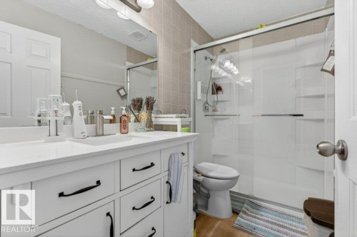 Bathroom featuring a white vanity with black hardware, a solid surface countertop, a large frameless mirror, a shower with a sliding glass door, and wood-finish flooring - 157 Kiniski Crescent, Edmonton, AB - Indoor Photo Showing Bathroom